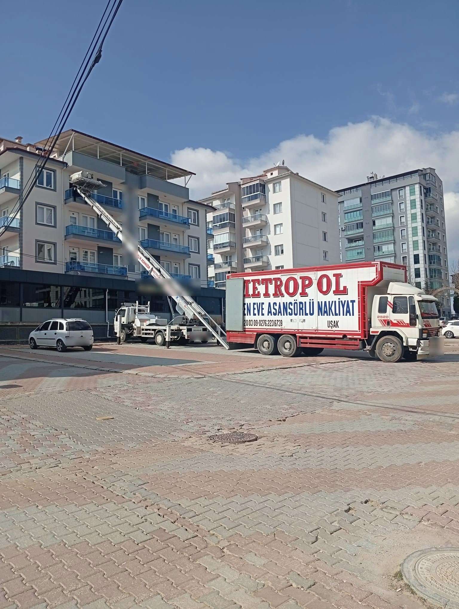 Mavi cam balkonlu bir apartmanın üst katına kurulu asansör ve karşı köşede bekleyen asansörlü nakliyat kamyonu