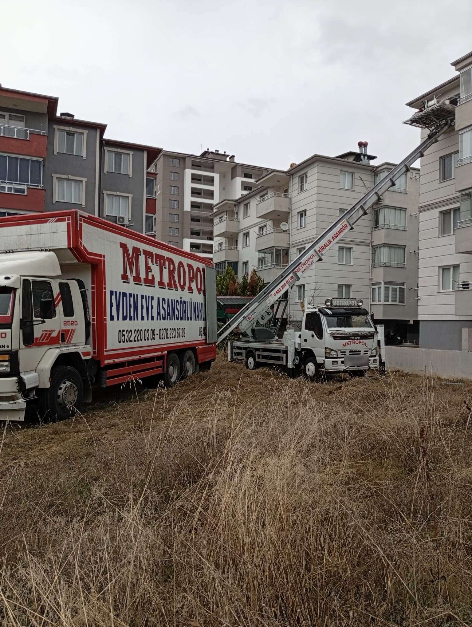 Boş bir arsanın kenarında kurulan asansörün platformu üst kat balkonunda, yanında Metropol kamyonu