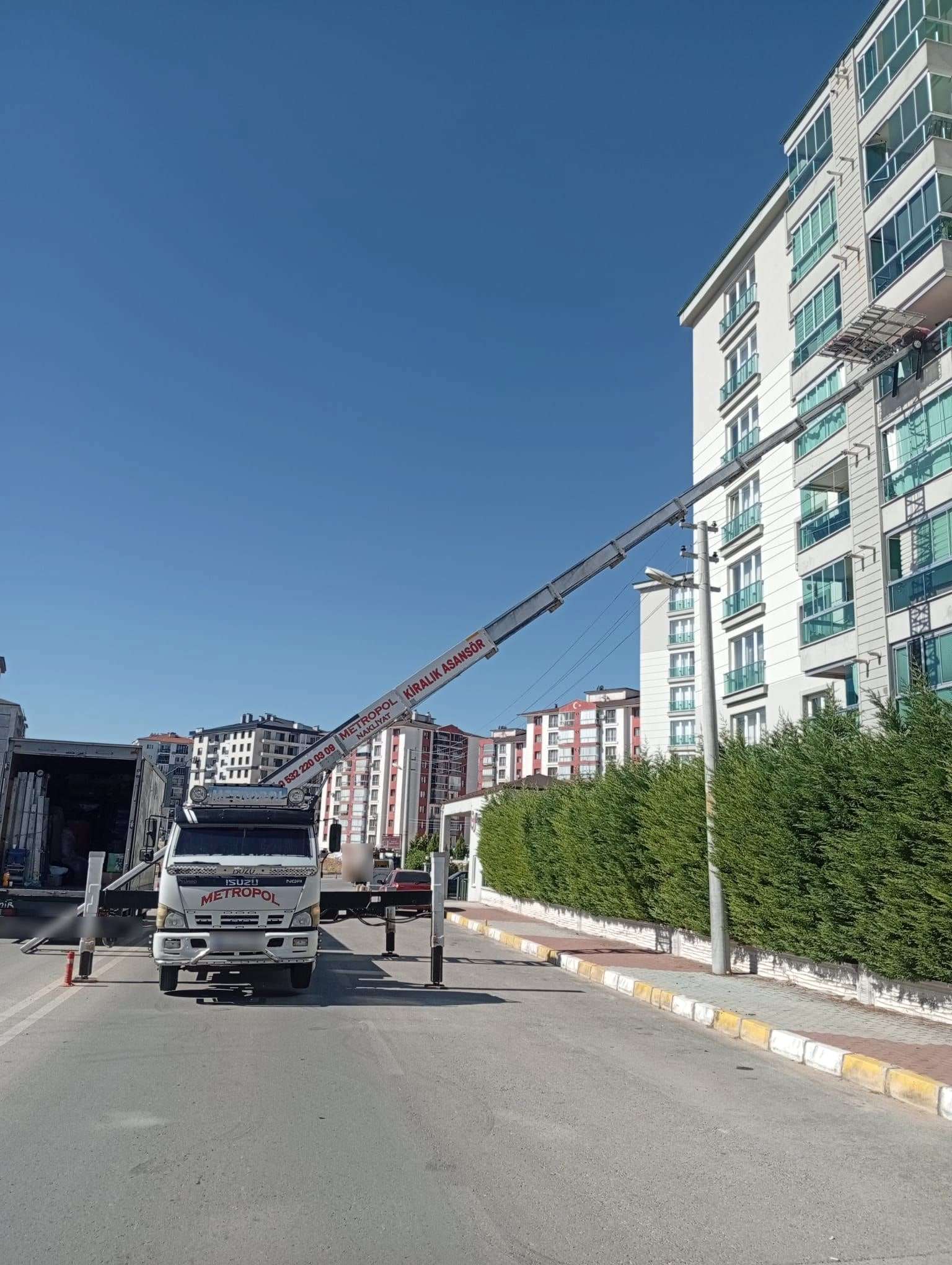 Uşak’ta çit boyunca uzanan bir caddede ayakları açılmış asansör aracı, platform yandaki binanın orta katlarında