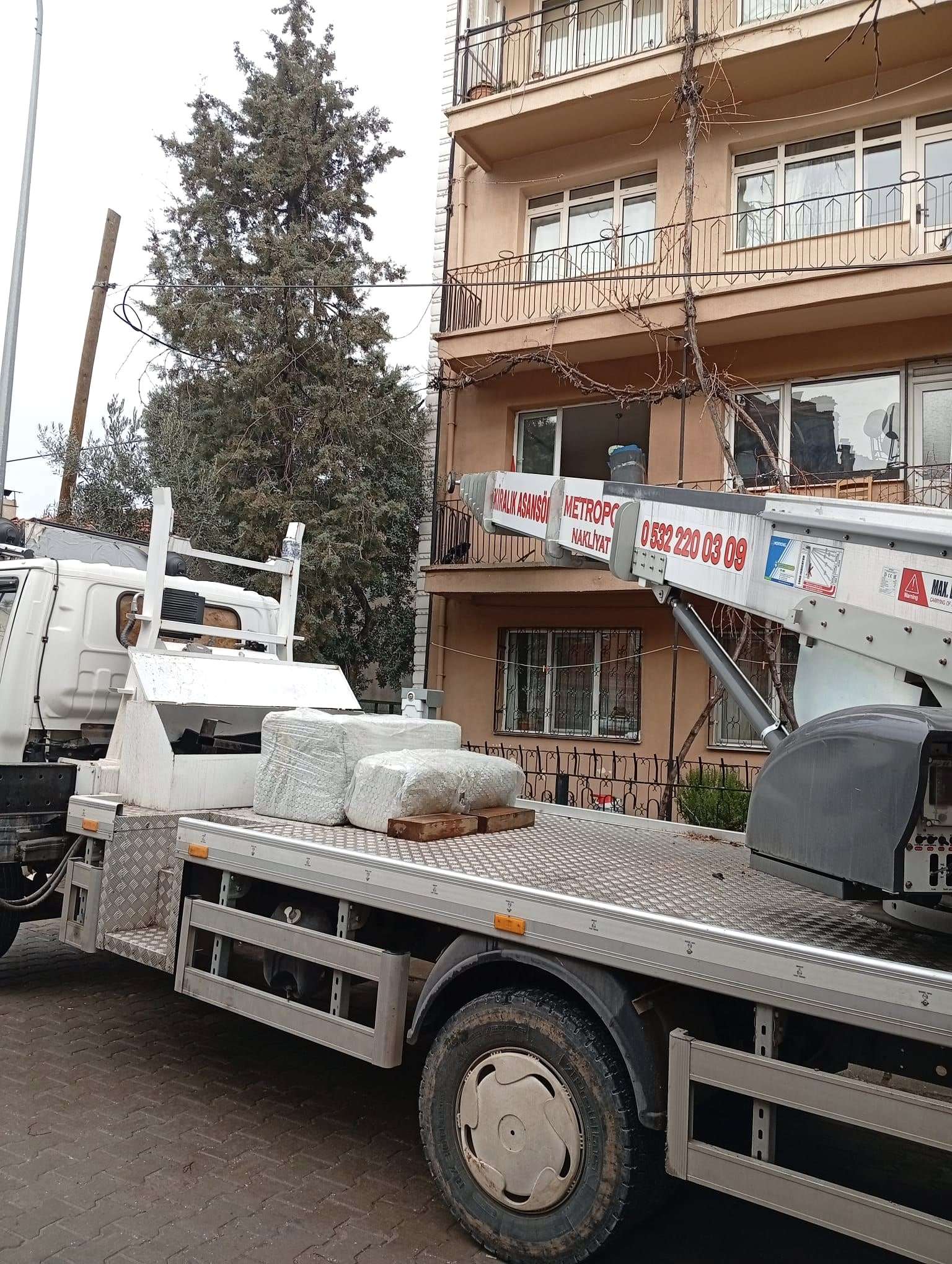Asansör takılı aracın kasasında streçlenmiş eşyalar; asansör kolu eski bir apartmanın balkonuna yönelmiş