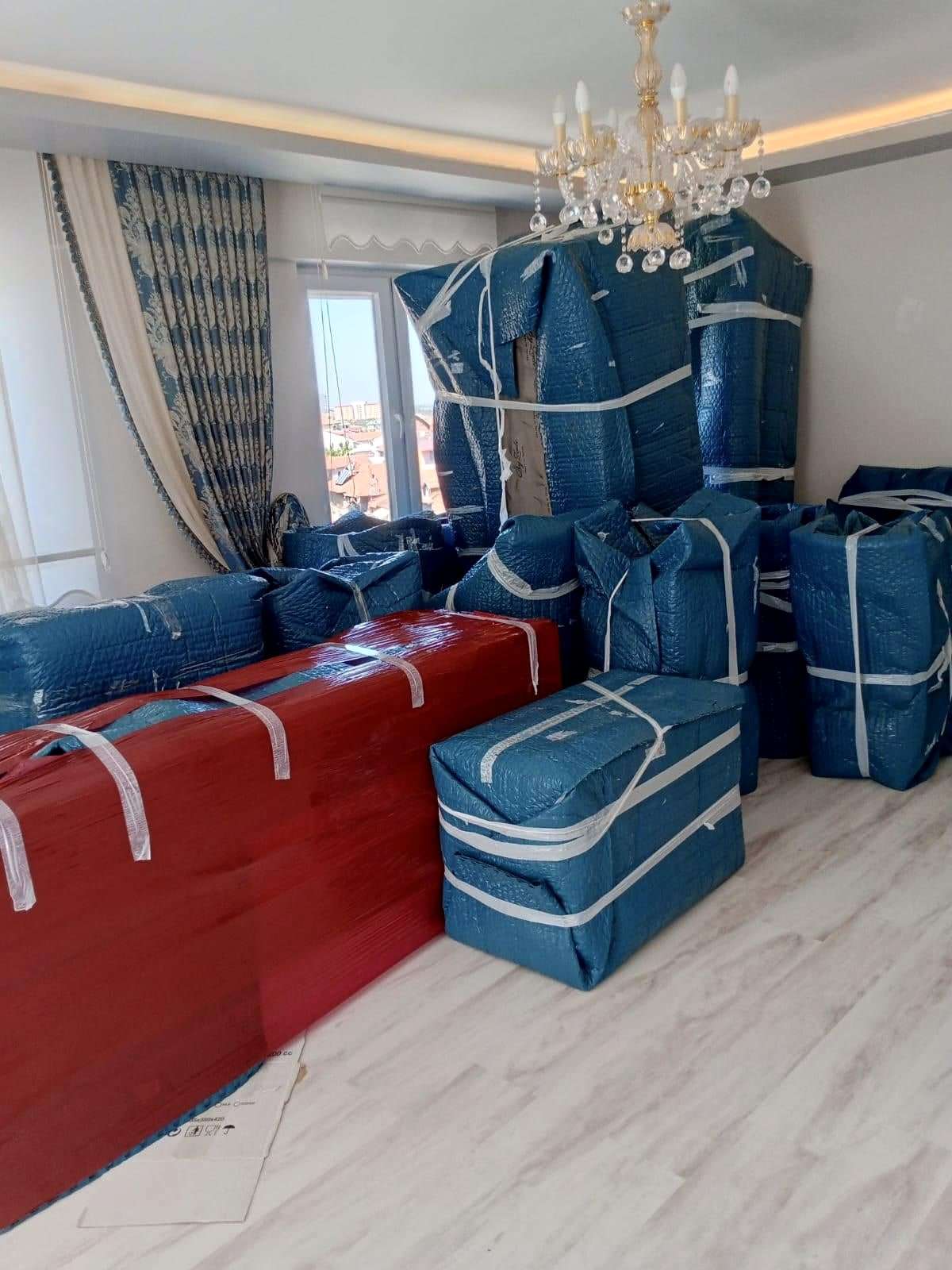 Avizeli bir salonda mavi ve kırmızı ambalajla paketlenip kayışla bağlanmış koltuklar ve eşyalar