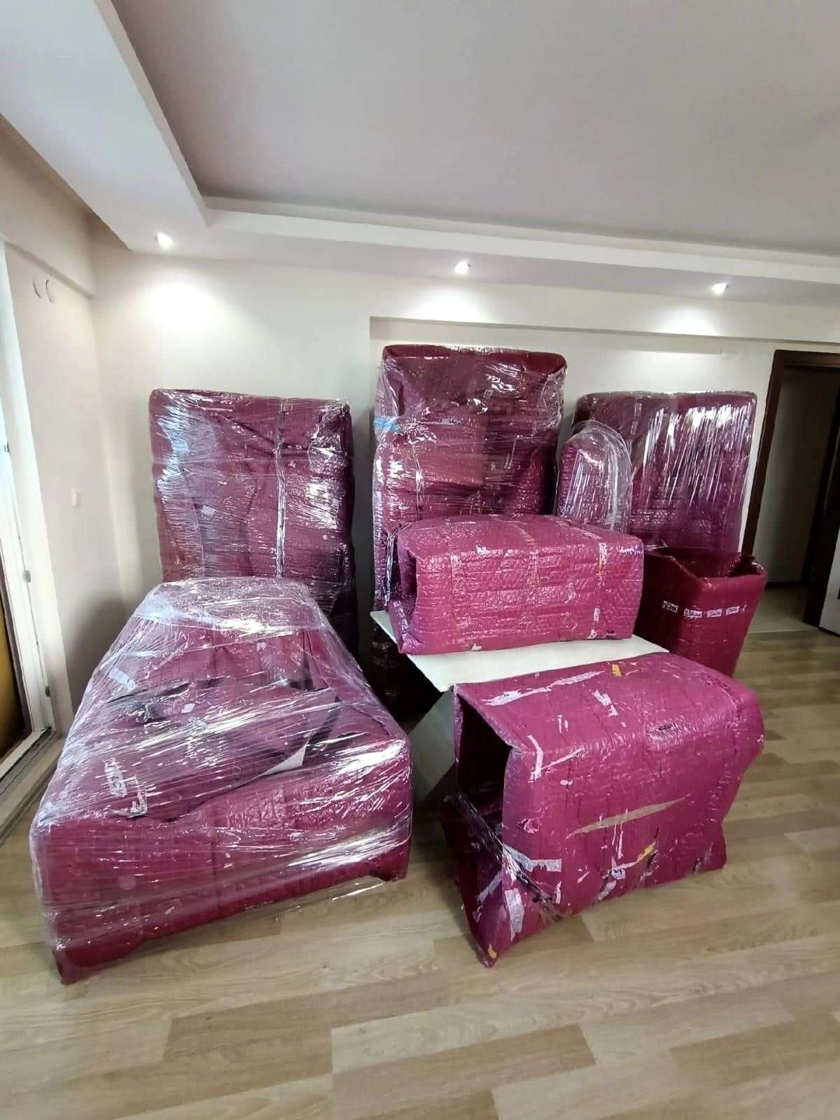 Bordo örtü ve streçle sarılmış koltuk takımı, üzerine paketli eşya konmuş beyaz masa ile salonda