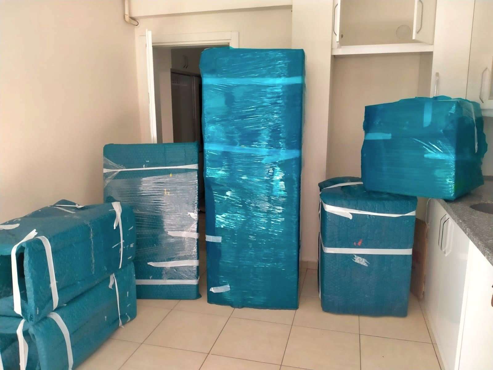 Mutfakta turkuaz ambalajla paketlenmiş buzdolabı, beyaz eşyalar ve bantlanmış koliler