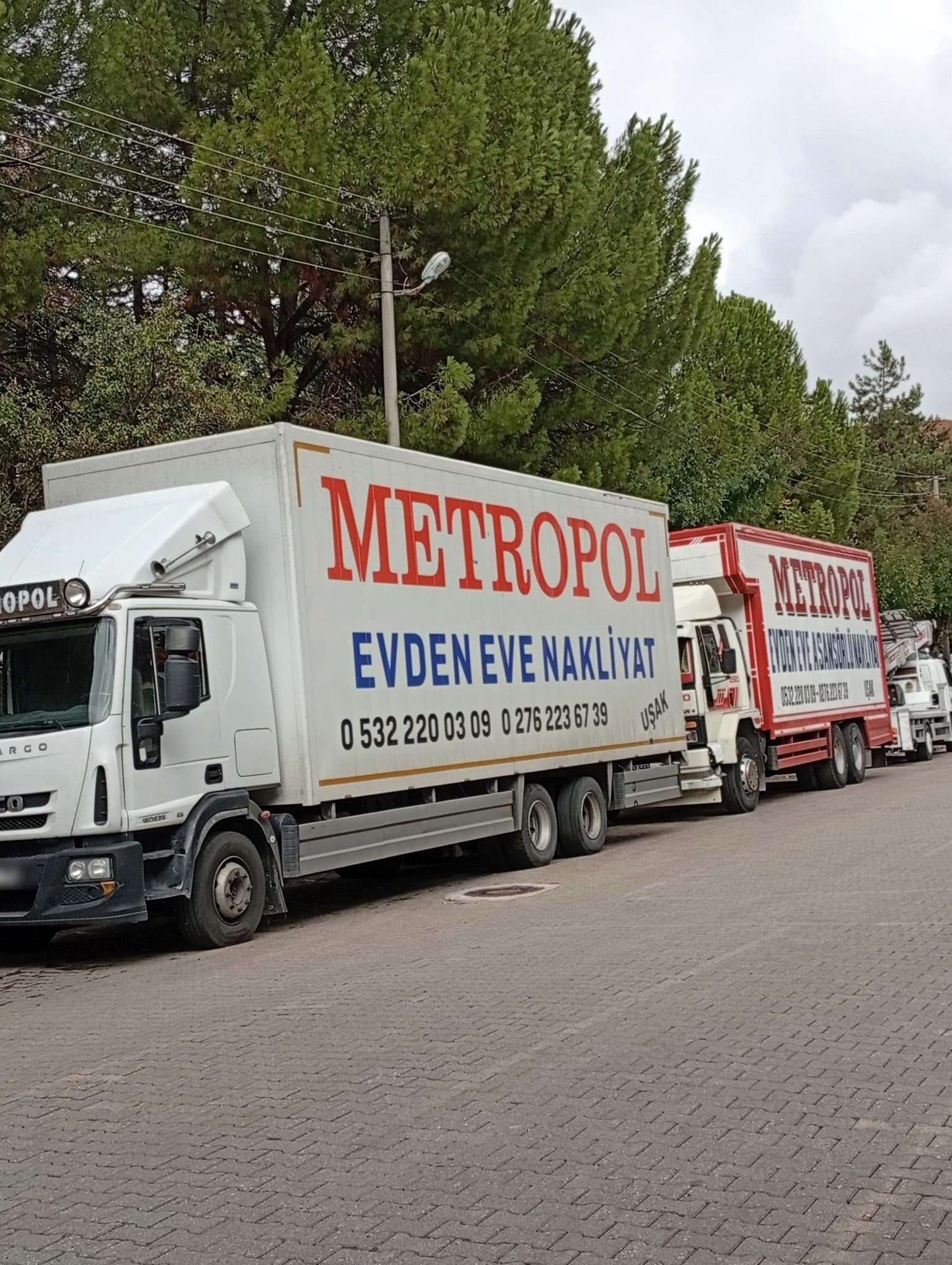 Ağaçlı bir yolda arka arkaya park etmiş Metropol Evden Eve Nakliyat kamyonları ve asansör aracı